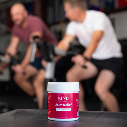 Container of KIND Joint Relief on a table with two people on stationary bikes in the background