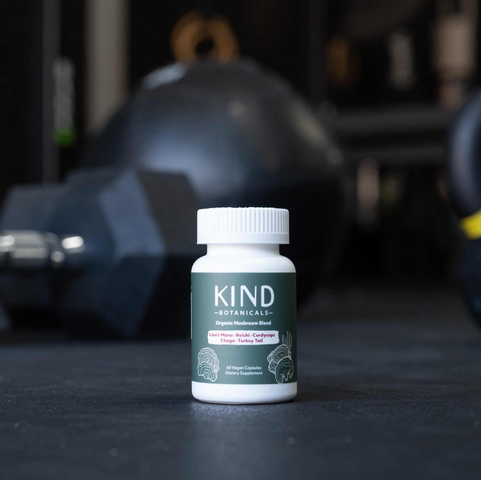 Bottle of Kind Botanicals supplement on a dark surface with gym equipment in the background