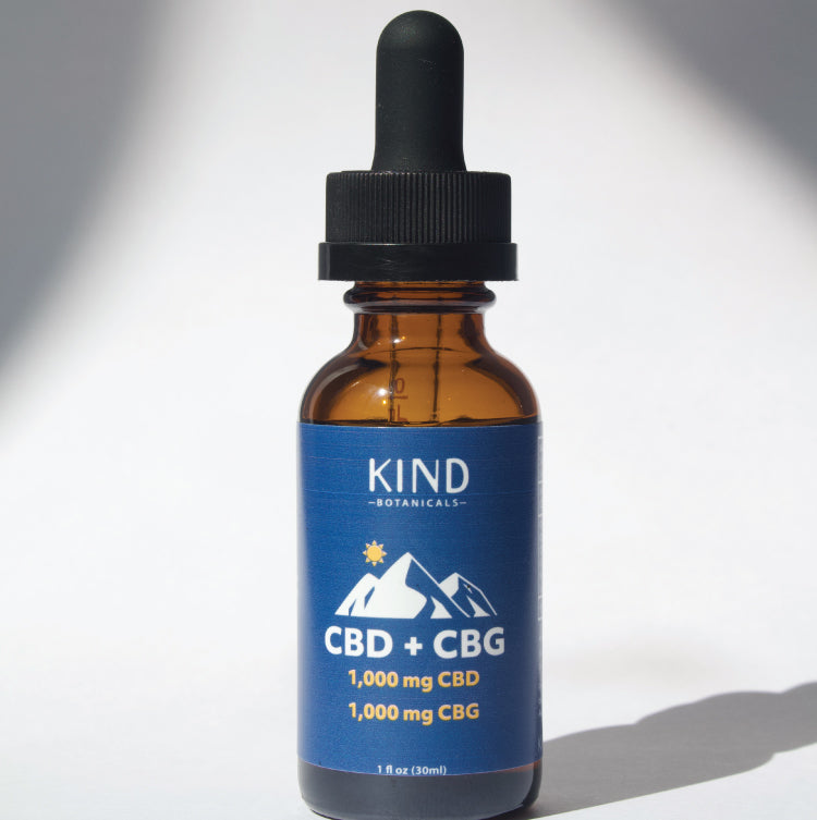 An amber glass dropper bottle of KIND Botanicals CBD + CBG tincture with a blue label and black dropper cap, photographed on a white background with soft shadows.